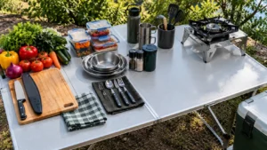 Bancada de alumínio para camping com vegetais frescos, tábua de corte, faca protegida, utensílios limpos, recipientes fechados e fogareiro desligado em suporte separado.