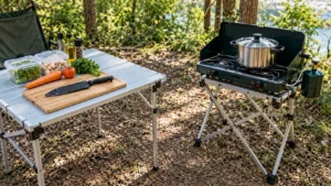 Cozinha de camping para duas pessoas com mesa dobrável cinza, tábua de corte, cenoura, tomate, faca protegida, recipientes limpos e fogareiro desligado em suporte separado.