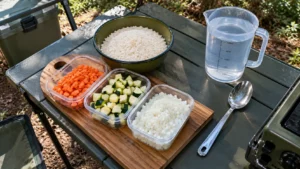 Bancada de camping com cenoura em cubos, abobrinha, cebola picada, arroz branco cru, recipiente graduado de água potável e colher metálica.