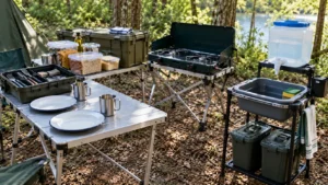 Mesa de camping com dois pratos reutilizáveis limpos, duas canecas metálicas, caixa com utensílios secos, bacia de lavagem, reservatório de água e fogareiro desligado ao fundo.
