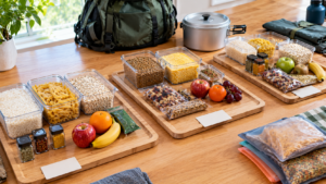 Bancada de cozinha com ingredientes de camping organizados por refeições, incluindo recipientes com arroz, macarrão, aveia, temperos, frutas inteiras e mochila ao fundo.