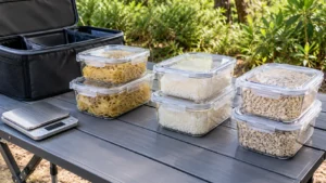 Mesa dobrável de camping com recipientes transparentes contendo porções de macarrão, arroz e aveia, caixa organizadora aberta e balança digital desligada.